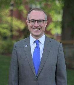 john maura smiling wearing a grey suit with a blue tie.